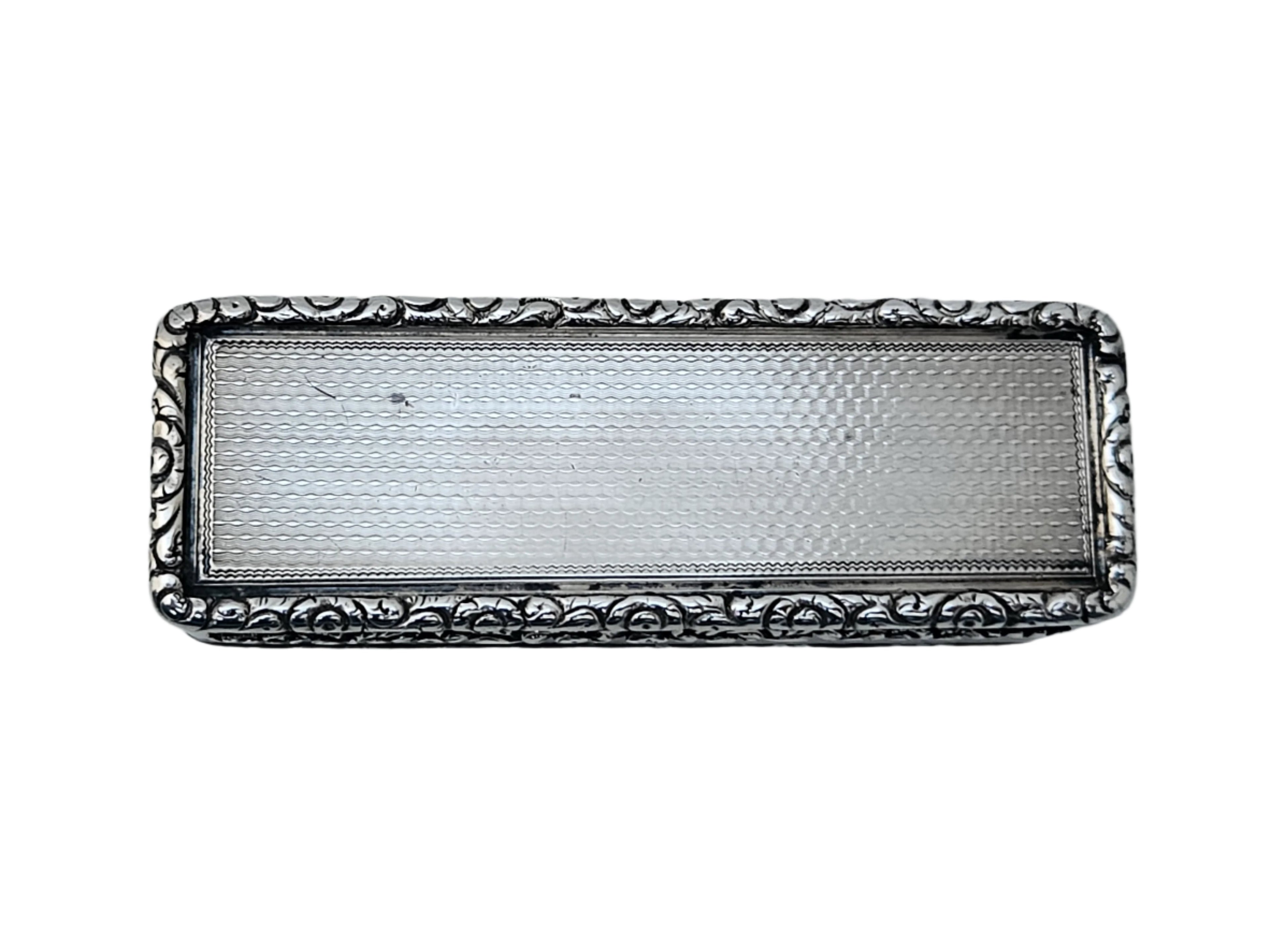 A STERLING SILVER 19TH CENTURY ANTIQUE ENGLISH SNUFF BOX by SEVENTH PLATEAU, featuring ornate floral patterns around its edges, rests on a marble surface. Its top is smooth with a textured center.