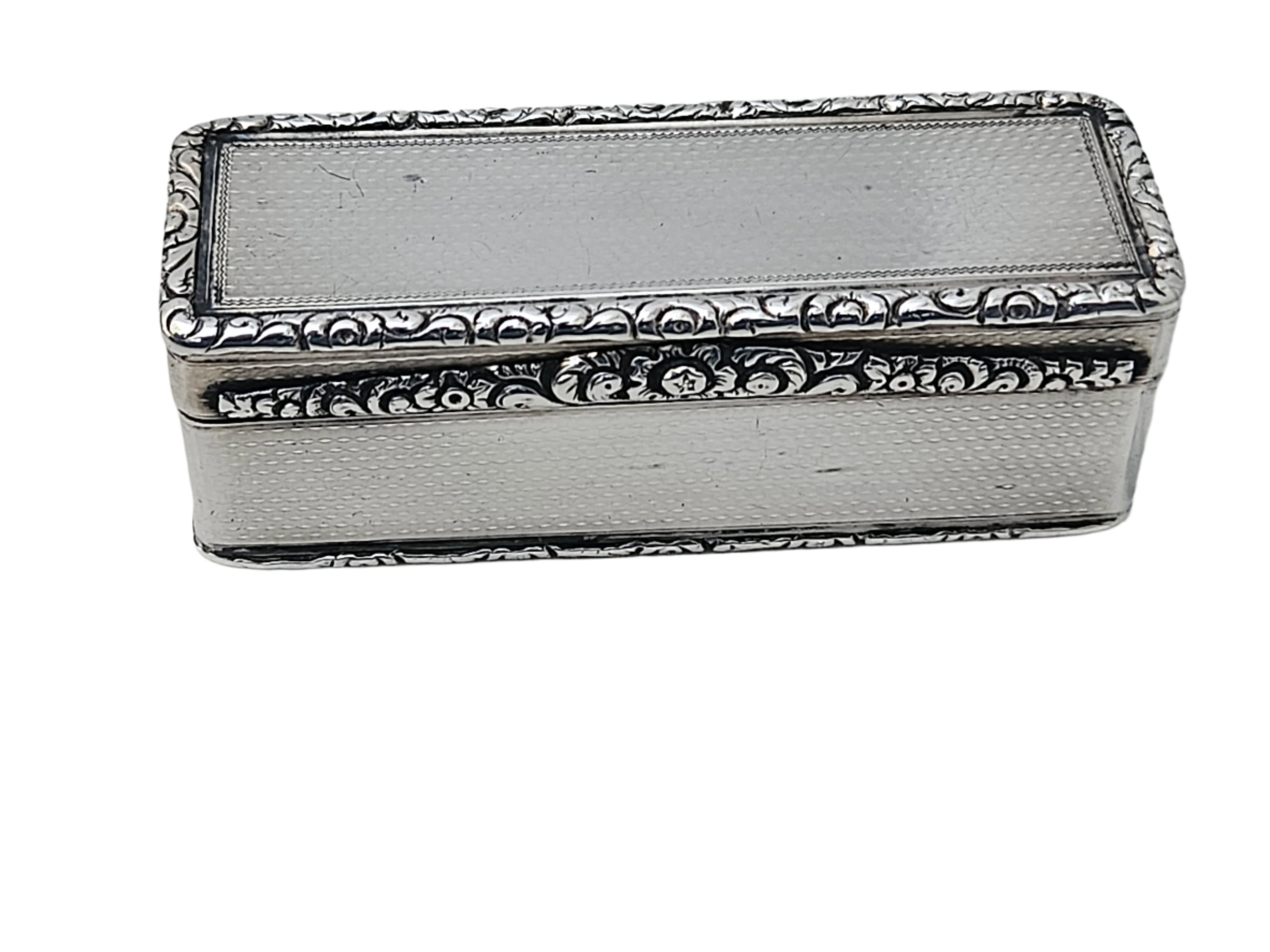 A rectangular STERLING SILVER 19TH CENTURY ANTIQUE ENGLISH SNUFF BOX by SEVENTH PLATEAU, featuring ornate floral engravings on its edges and a textured surface, placed on a light marble surface.
