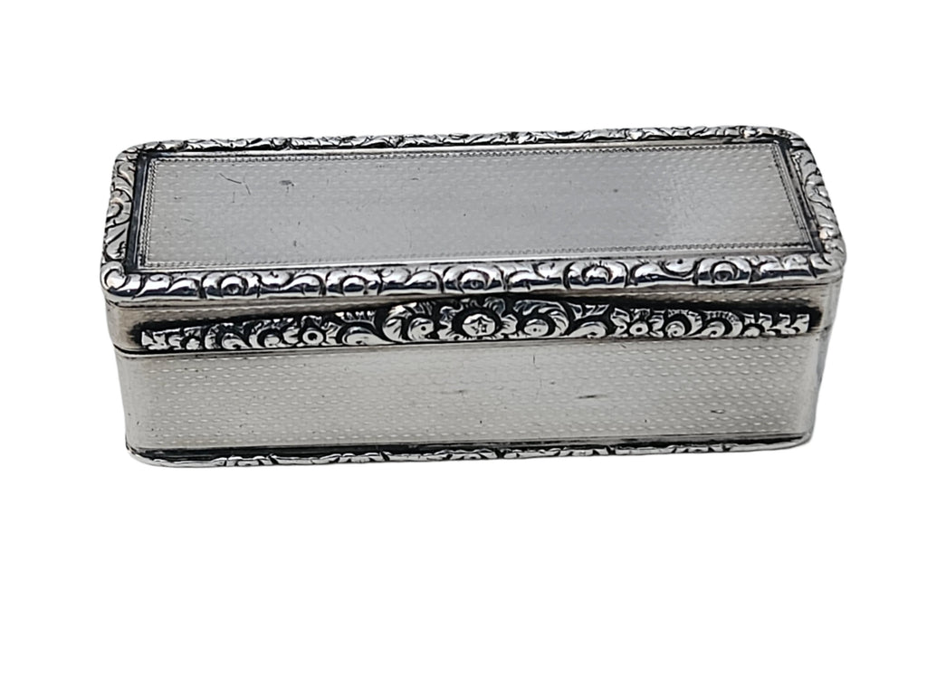 On a white marble surface rests the SEVENTH PLATEAU Sterling Silver 19th Century Antique English Snuff Box, featuring ornate floral engravings on the edges and a smooth, plain center on its top.