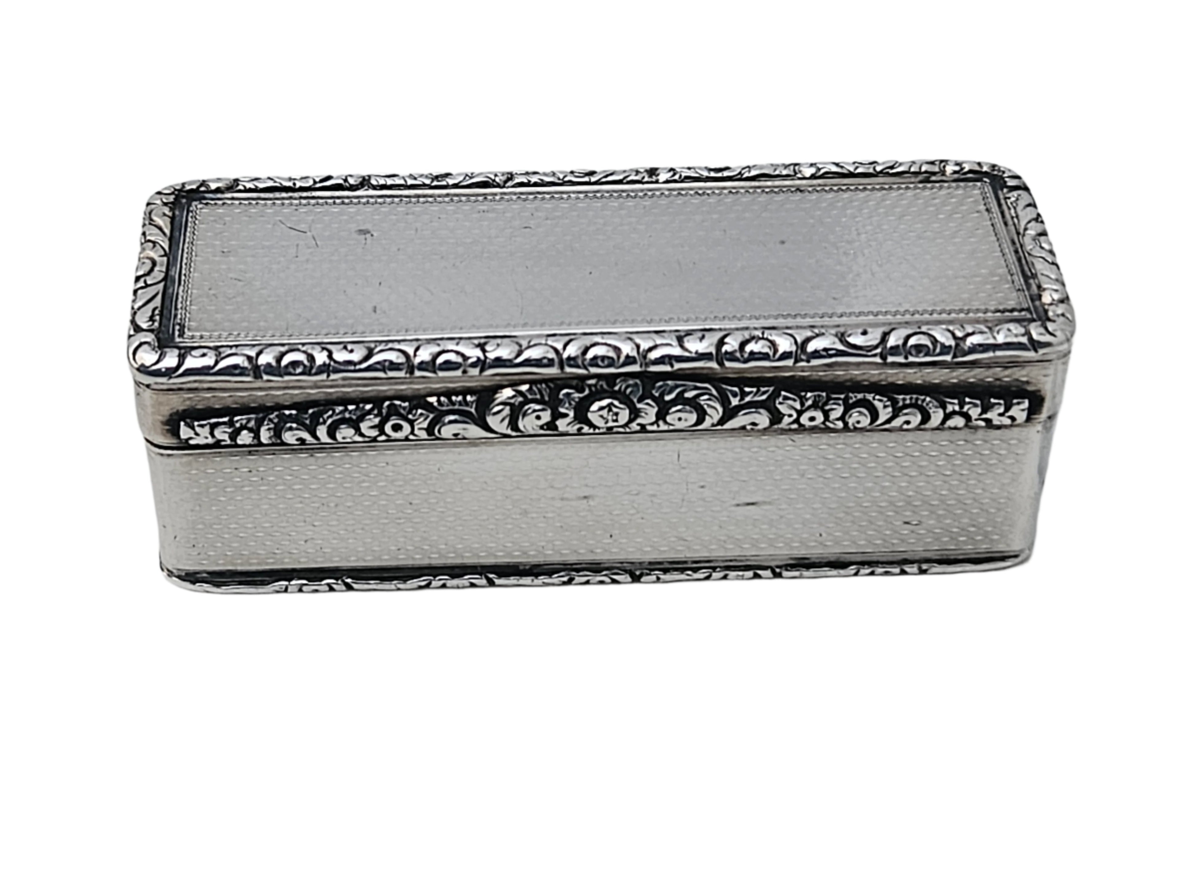 On a white marble surface rests the SEVENTH PLATEAU Sterling Silver 19th Century Antique English Snuff Box, featuring ornate floral engravings on the edges and a smooth, plain center on its top.